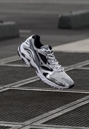 Weißer Sportschuh aus Mesh- und Synthetikmaterial, mit schwarzem Akzent, abgerundeter Spitze und gepolsterter Sohle, über einer Gitterfläche positioniert.