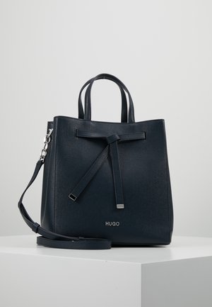 Sac à main HUGO en cuir noir texturé avec double poignées, un détail de nœud à l'avant et une bandoulière amovible sur une surface blanche.