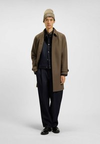 Tan uldecoat over en mørk denimjakke, parret med løse mørke bukser og sorte sko. En beige strikket beanie fuldender outfittet.