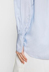 Camicia azzurro chiaro a maniche lunghe con righe sottili, dotata di polsini con bottoni e fessura laterale. Tessuto liscio e vestibilità sartoriale.