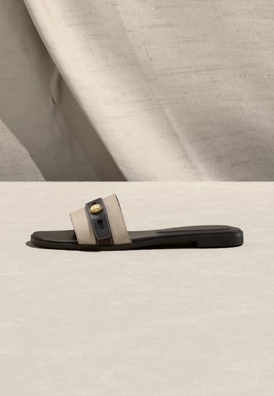 Πλατφόρμα σανδάλι με ιμάντα από μπεζ υφή υφάσματος, μαύρη σόλα και χρυσό στρογγυλό έμβλημα στον ιμάντα, τοποθετημένο σε ουδέτερο υφασμάτινο φόντο.