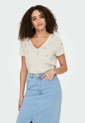 T-shirt crème à manches courtes en V avec des motifs en feuilles d'or, associé à une jupe crayon en denim bleu avec une fente avant et des poches.