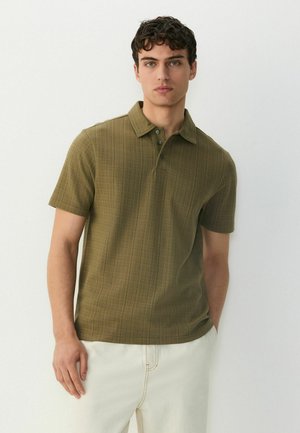 Camisa polo de manga corta en verde olivo, con un patrón texturizado, cuello y tapeta de tres botones; combinada con pantalones blancos.