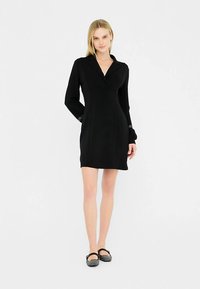 Must kleit, millel on krae, pikad varrukad ja keha järgiv siluett. Sile kangas minimaalsete detailidega. Kombineeritud mustade madalate kingadega.