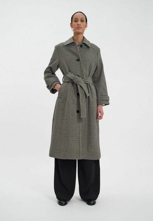 Manteau long à carreaux noir et blanc, avec une taille ceinturée, de grands boutons et un col classique, associé à un pantalon noir à jambes larges.