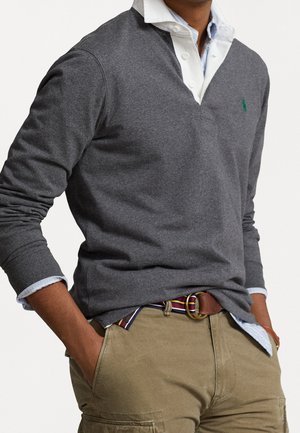 Mężczyzna w szarej koszulce rugby z długim rękawem na białej koszuli z kołnierzykiem, w paskowanym pasku i beżowych spodniach, z rękami w kieszeniach.