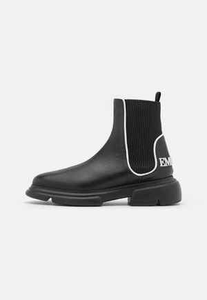 Bota tobillera de cuero negro con suela gruesa, panel lateral elástico acanalado delineado en blanco y texto blanco parcial en el talón.