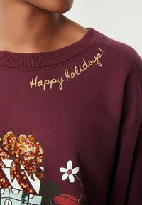 Sweatshirt bordeaux avec broderie "Joyeuses fêtes !" en fil d'or. Présente un design de cadeau en sequins avec un accent floral et un col côtelé.