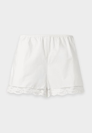 Kvinde med kort mørkt hår iført hvid ærmeløs top med blonderkant og shorts, stående med den ene hånd i lommen og sorte flade sko med nitter.