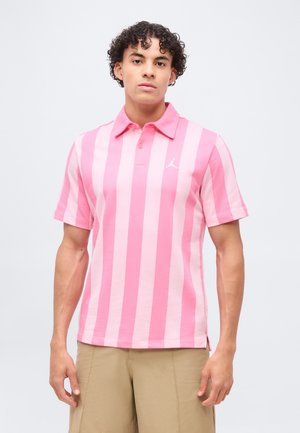 Jonge man met krullend haar die een roze gestreept poloshirt met korte mouwen en beige broek draagt tegen een effen achtergrond.