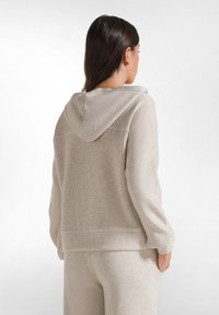 Beige hoodie med ett mjukt, texturerat tyg som har en subtil glans. Inkluderar en huva och sänkta axlar, visad från baksidan.