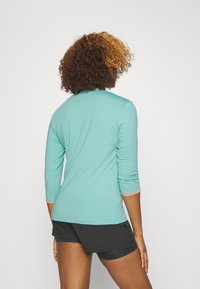 Türkises Langarmshirt mit 3/4-Ärmeln, versehen mit orangefarbenen Akzenten an den Bündchen, kombiniert mit schwarzen Shorts. Weicher, leichter Stoff.