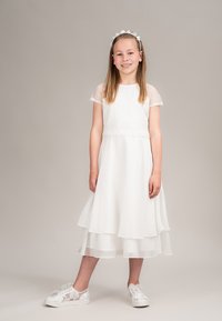 Weißes Kleid mit kurzen Spitzenärmeln, mehrlagigem Rockdesign und perlenverziertem Taillenbereich. Kombiniert mit transparenten Spitzen-Sneakern mit Schleifenakzenten.