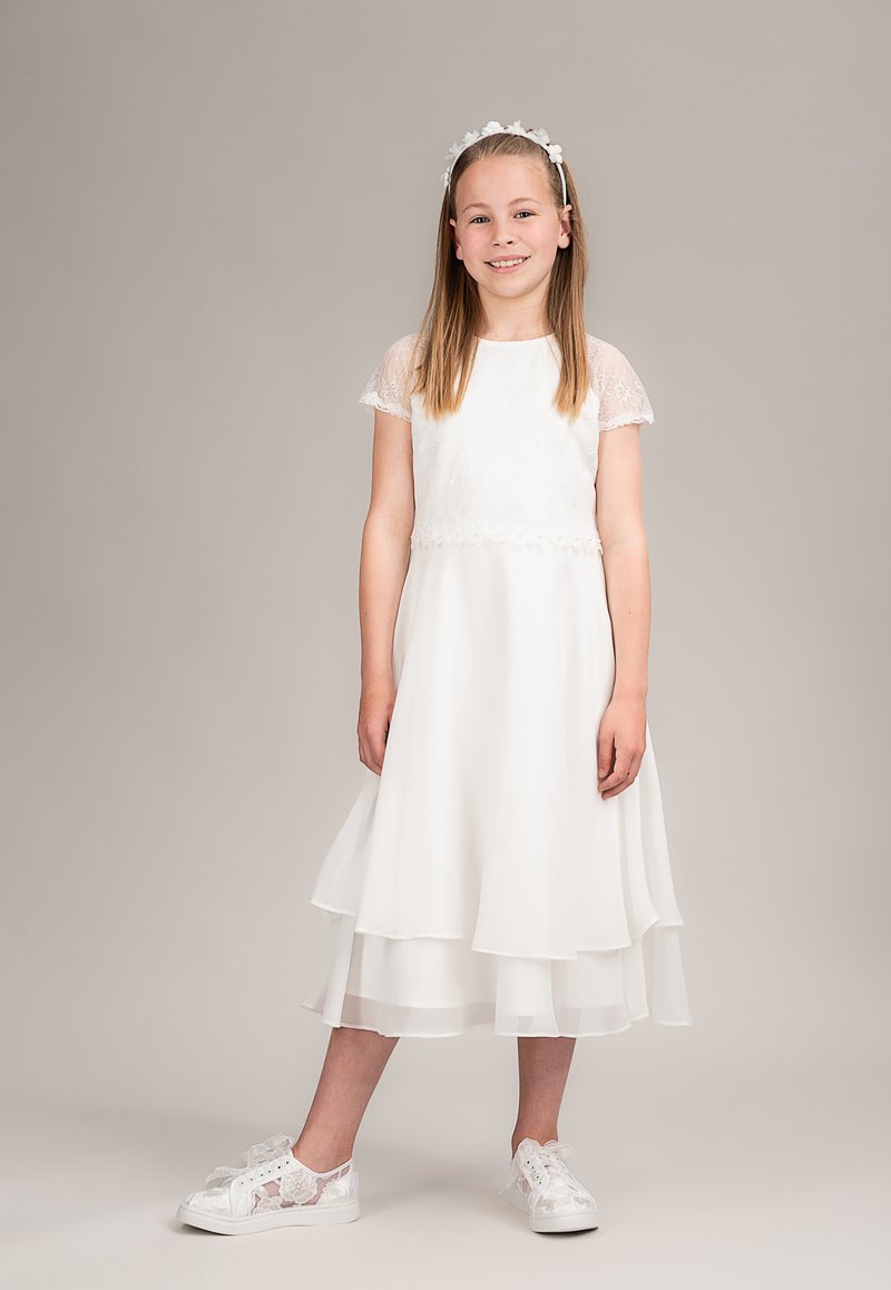 Weißes Kleid mit kurzen Spitzenärmeln, mehrlagigem Rockdesign und perlenverziertem Taillenbereich. Kombiniert mit transparenten Spitzen-Sneakern mit Schleifenakzenten.