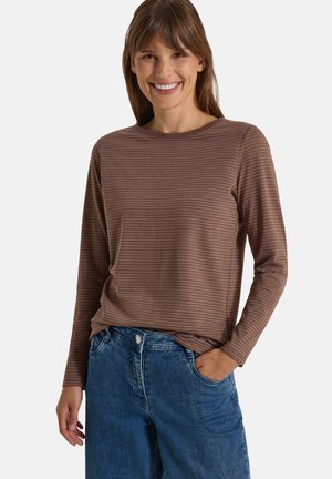 Kvinde smilende iført brun langærmet stribet skjorte og blå jeans med den ene hånd i lommen mod ensfarvet baggrund.