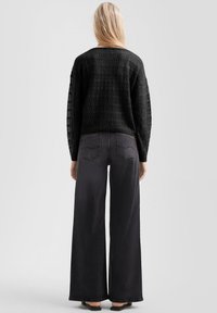Blonde vrouw die zich met de rug naar de camera draait, gekleed in een zwart gehaakt shirt met lange mouwen en hoge taille brede zwarte jeans, met sandalen op een lichte achtergrond.