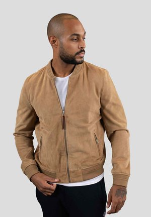 Veste bomber en suede tan avec des poignets et un ourlet côtelés, dotée d'une fermeture éclair à l'avant et de deux poches latérales. Portée sur une chemise blanche.