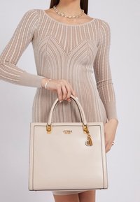 Beige gestructureerde handtas met goudkleurige hardware, bovenhandvatten, logo-detail en een aanhangsel. De gladde textuur contrasteert met de ribbelstof.