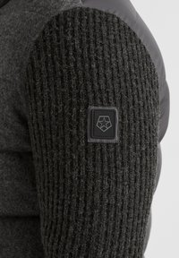 Gestickter schwarzer Stoff am Ärmel mit strukturiertem Muster, ergänzt durch einen grauen gepolsterten Bereich. Besitzt ein kleines schwarzes Logo-Patch.