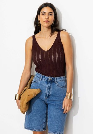 Femme aux longs cheveux foncés portant un haut en maille marron sans manches, un short en jean bleu, des bracelets en or et tenant un sac beige.