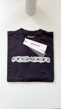 Črna majica s krožnim izrezom, ki vsebuje sijajni logo "PEÑAGADOR" v srebrni barvi. Vključuje belo oznako z navodili za nego.