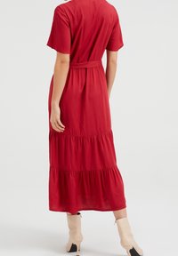 Červené maxi šaty s krátkými rukávy, páskem na zavázání, vrstveným spodním lemem a texturovanou tkaninou. Nosené s nahými kozačkami po kolena.