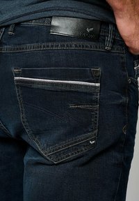 Tmavě modré džínové kalhoty mají výraznou zadní kapsu s bílým zipem, kontrastní prošívání a černou koženou štítek na pásku.