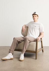 White cotton t-shirt with small text, beige sweatpants, white sneakers with tan soles, seated in a wooden armchair with a fabric seat.