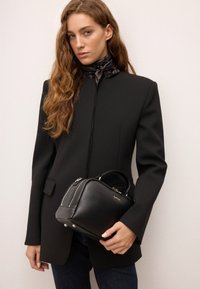 Črna usnjena torbica z oblikovano strukturo, srebrnimi dodatki in zgornjim ročajem. Nosila jo je manekenka v črnem blazerju, ki prikazuje gladko teksturo.