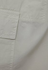 Tissu beige clair avec des coutures doubles visibles et des patchs rectangulaires superposés sur une surface textile.