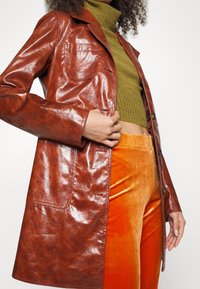 Manteau en cuir brillant marron avec un col, des poches avant et une fermeture à boutons, porté sur un pull en maille vert et un pantalon en velours orange.