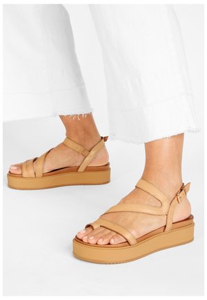 Ben klædt i hvide cropped bukser og brune platformsandaler med ankelremme, som står på en hvid overflade.