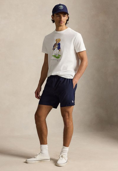 Νεαρός άνδρας που φοράει λευκό T-shirt με σχέδιο αρκούδας, σκούρο μπλε σορτς, λευκά αθλητικά παπούτσια και σκούρο μπλε καπέλο μπέιζμπολ, στέκεται με το ένα χέρι στην τσέπη.