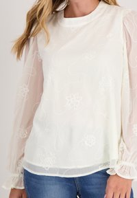 Witte blouse met doorschijnende, opgezette lange mouwen. Bevat bloemgebruikste borduursels en draaiende patronen, textuurstof en een ronde halslijn.