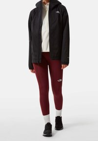 Person, der eine schwarze Kapuzenjacke, einen cremefarbenen Halbzip-Pullover, bordeauxfarbene Leggings, weiße Socken und schwarze Sportschuhe trägt, steht vor einem weißen Hintergrund.