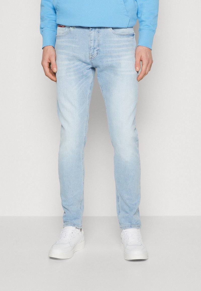 Jeans de mezclilla azul claro con un corte slim, desvanecimiento sutil y diseño de cinco bolsillos. Combinados con zapatillas deportivas blancas y una sudadera azul.