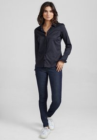 Zwarte, langwerpige knopshirt met kraag, slim fit ontwerp en gerimpelde manchetten, gecombineerd met donkerblauwe skinny jeans en witte sneakers.