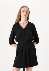 Schwarzes Kleid mit V-Ausschnitt, drei Viertelärmeln und tailliertem Bund. Der Stoff wirkt glatt mit einem leichten Glanz und ist für eine lockere Passform konzipiert.