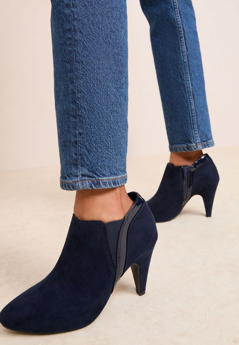 Persona che indossa stivaletti alla caviglia in camoscio blu con tacco alto e cerniera laterale abbinati a jeans in denim blu su sfondo semplice.