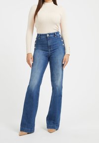 Jeans z wysokim stanem i rozszerzanymi nogawkami w średnim niebieskim denimu z złotymi guzikami. Gładka textura i niewielkie przetarcia na kolanach, zestawione z dopasowanym beżowym topem.