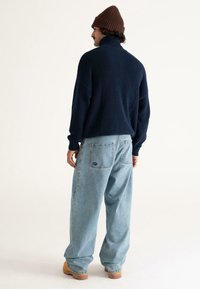 Man wearing a brown knit beanie, navy blue sweater, loose light blue jeans, and tan boots, standing facing away on a plain white background.