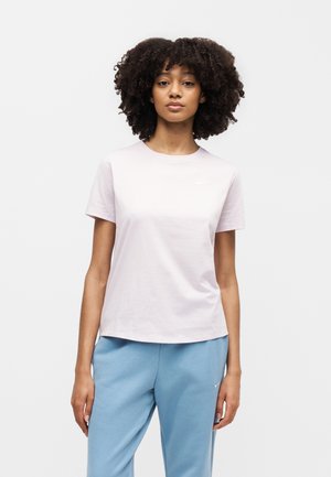 Femme aux cheveux bouclés portant un t-shirt Nike rose clair et un pantalon de survêtement Nike bleu clair, debout devant un fond blanc uni.