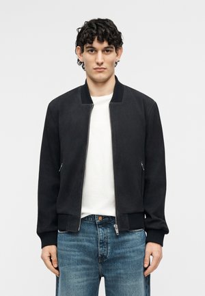 Νεαρός άνδρας που φοράει μαύρο bomber μπουφάν με φερμουάρ, λευκό μπλουζάκι και μπλε τζιν, όρθιος μπροστά σε απλό φωτεινό φόντο.