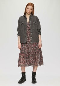 Chemise en denim grise sur une robe noire à fleurs avec une jupe à volants. Le mannequin porte des bottes noires à lacets. Fond blanc uni.