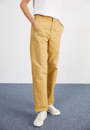 Person iført beige bukser med lige ben, hvide sneakers og en hvid skjorte, stående med hænderne i lommerne på et blåt tæppe.