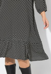 Schwarzes Kleid mit weißem Herzmuster, gerafftem Saum und lockeren Ärmeln. Getragen mit knielangen schwarzen Stiefeln. Glatter Stofftextur.