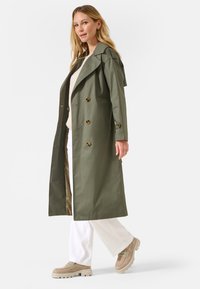 Trench-coat vert olive avec un design croisé, de grands boutons et une capuche, associé à un pantalon blanc et des chaussures à plateforme beiges.