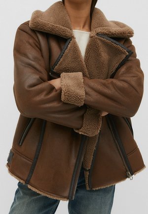 Person iført en brun shearling-jakke med foldede arme, der viser en blød tekstureret krave og manchetter over en lys sweater og blå jeans.