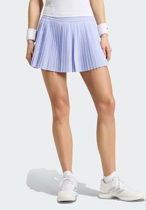Mujer que lleva una falda de tenis plisada de color lila claro, camiseta sin mangas blanca, muñequeras blancas y zapatillas deportivas blancas, de pie frente a un fondo liso.
