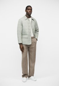 Homme portant une veste matelassée gris clair, un t-shirt blanc, un pantalon large beige et des baskets blanches, debout devant un fond uni.